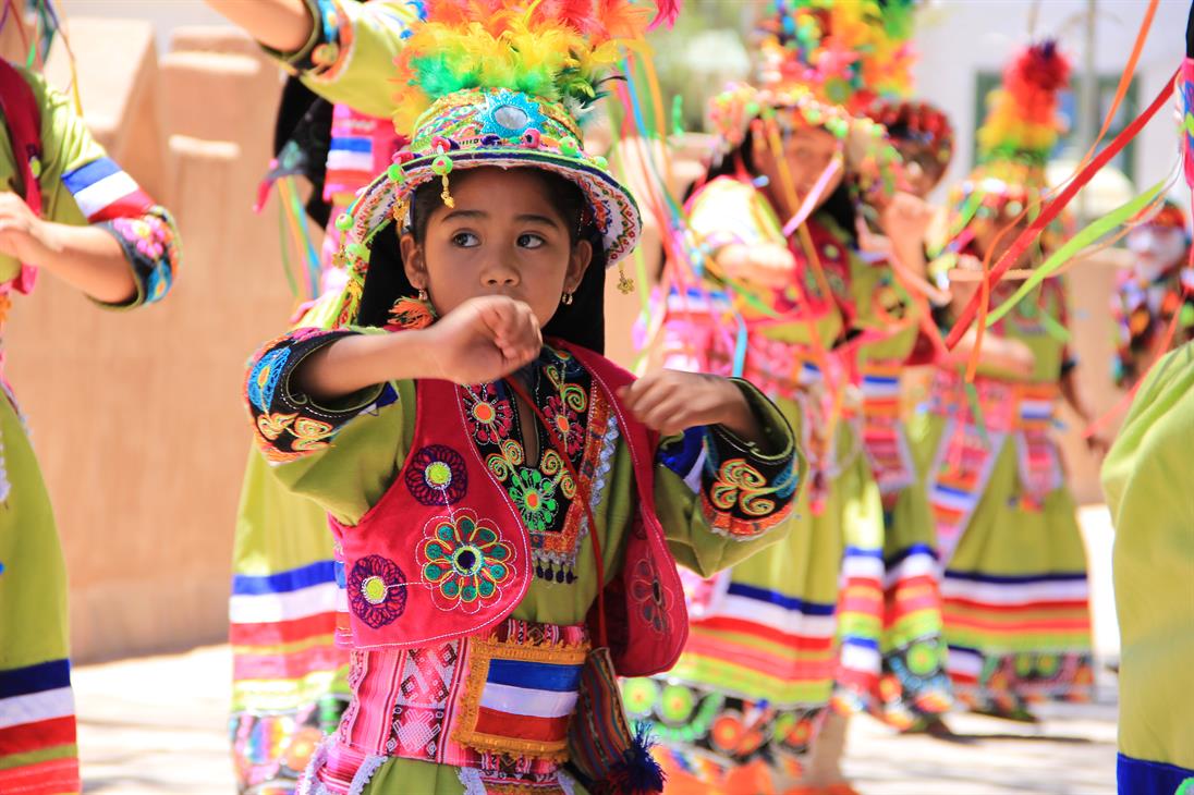 Ein junges Mädchen im traditionellen Kostüm tanzt gemeinsam mit einer Gruppe älterer Tänzerinnen bei einem Straßenfest im peruanischen San Pedro de Atacama.